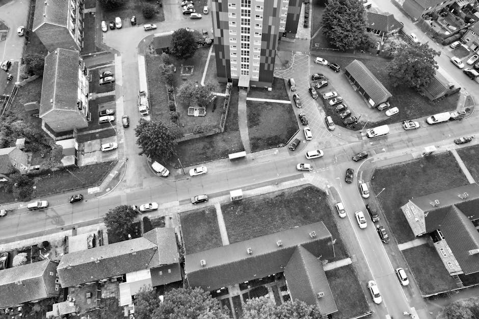 An aerial black-and-white photograph showing a residential area with multiple houses and apartment buildings separated by roads, pathways, and small green spaces with trees. Several cars are parked along the streets and in driveways, with some vehicles actively parked or moving at intersections. In the foreground, there is a row of terraced houses with tiled roofs, and behind them, larger apartment blocks with multiple floors and windows. The scene captures a typical urban neighbourhood layout, with vehicles indicating loading or unloading activities related to house removals or home relocation services. This visual reflects the kind of environment where [COMPANY_NAME], such as Man With a Van Barking, might facilitate furniture transport, packing and moving, or logistics involving vans and other equipment for effective transport and relocation, aligned with local council rules for permits, as indicated by the page title.