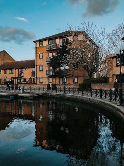 View of a multi-storey brick residential building situated near a body of water, with a paved walkway and black metal railing along the waterfront. Several people are walking along the pavement, and a large leafless tree stands in front of the building. The image captures a scene during daylight hours with a partly cloudy sky. The photograph is relevant to house removals and relocating services, illustrating a typical street environment that could be involved in a local house move or furniture transport in Barking Riverside, as described in the Barking Riverside moving guide on manwithavanbarking.co.uk. The scene emphasizes urban residential surroundings suitable for loading and moving activities, with visible housing, outdoor public space, and a calm water feature contributing to the context of local home relocations.
