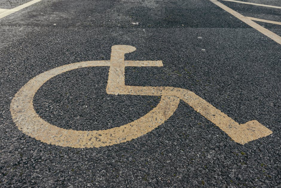 A close-up view of a designated accessible parking space in an asphalt driveway, featuring a yellow wheelchair symbol painted onto the surface. The painted lines are slightly worn, with some chipping and weathering visible. The surface is textured with small stones and rough patches. In the background, faint white parking bay lines are visible, indicating that the area is part of a parking lot or designated drop-off zone. This parking space is empty, with no vehicles or other objects present. Occasionally, Man With a Van Barking's removals team may use such accessible parking spots for loading and unloading household items during moving or home relocation services, aligning with transportation logistics and packing processes associated with house removals as outlined in the Barking Riverside moving guide.