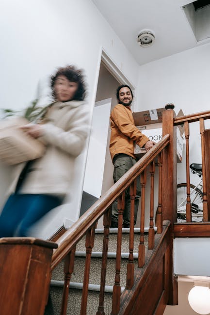 A man with a beard and smiling face stands on a carpeted staircase inside a home, holding a cardboard box labeled 'cook' and other boxes behind him. The staircase has wooden handrails and balusters, l