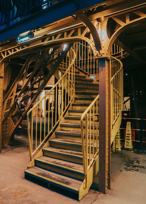 A man with a beard and smiling face stands on a carpeted staircase inside a home, holding a cardboard box labeled 'cook' and other boxes behind him. The staircase has wooden handrails and balusters, l