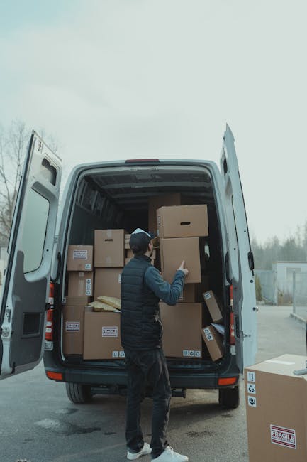 A person wearing a dark vest, black trousers, and a cap is standing next to an open delivery van on a paved surface, engaged in the home relocation process. Inside the van, which is parked outdoors with a cloudy sky above, numerous cardboard boxes of various sizes are stacked, some sealed with packing tape and others partially open, revealing packaging materials such as bubble wrap and foam. The boxes are positioned on different levels within the van, indicating an organized loading or unloading process typical of professional removals. Several boxes with labels or barcodes are visible, suggesting inventory management during the move. Outside the van, additional boxes, including one marked 'fragile,' rest on the ground or are in the process of being loaded or unloaded, with the person using hand gestures to guide or communicate regarding the transport of these items. The environment appears to be a commercial or residential area, fitting for professional house or furniture removal services by Man With a Van Barking, where packing and loading operations are underway to facilitate efficient home relocation and furniture transport.