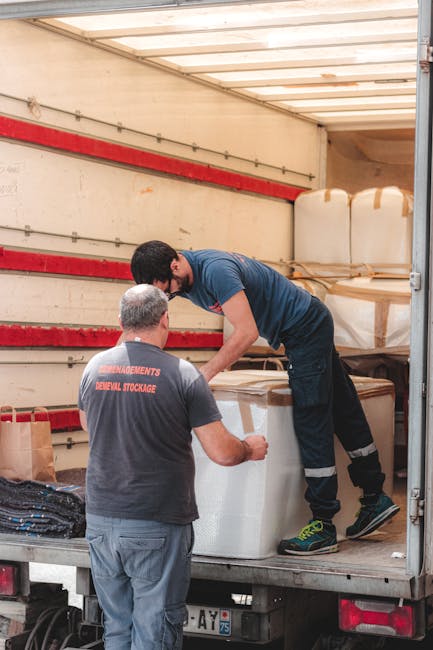 A person wearing a dark vest, black trousers, and a cap is standing next to an open delivery van on a paved surface, engaged in the home relocation process. Inside the van, which is parked outdoors with a cloudy sky above, numerous cardboard boxes of various sizes are stacked, some sealed with packing tape and others partially open, revealing packaging materials such as bubble wrap and foam. The boxes are positioned on different levels within the van, indicating an organized loading or unloading process typical of professional removals. Several boxes with labels or barcodes are visible, suggesting inventory management during the move. Outside the van, additional boxes, including one marked 'fragile,' rest on the ground or are in the process of being loaded or unloaded, with the person using hand gestures to guide or communicate regarding the transport of these items. The environment appears to be a commercial or residential area, fitting for professional house or furniture removal services by Man With a Van Barking, where packing and loading operations are underway to facilitate efficient home relocation and furniture transport.