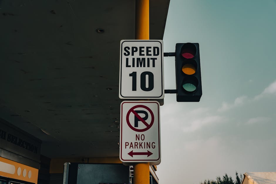 Close-up view of a tall yellow pole supporting a traffic light and two road signs under an overhanging structure. The upper sign displays a black and white speed limit of 10 miles per hour, indicating a low-speed zone likely related to residential or moving areas. Below it, a white sign with black and red markings shows a 'No Parking' restriction with a red circle and slash over a letter 'P', accompanied by a left-pointing arrow. The background reveals a partly cloudy sky with a hint of sunlight. The surrounding environment suggests a street or parking area adjacent to a building or service entrance, with visible roof edges and exterior walls. This setup is typical for areas requiring regulated parking and traffic control during home relocations or moving operations, as managed by companies like Man With a Van Barking, especially when securing parking suspensions or complying with council regulations for furniture transport and home removals.