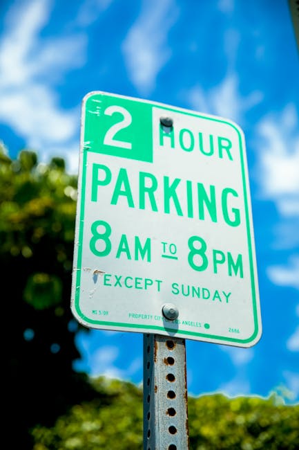 A green and white parking restriction sign mounted on a metal pole outdoors, indicating a two-hour parking limit from 8 am to 8 pm, Monday to Saturday, with an exception for Sundays. The sign is set against a background of blue sky with some clouds and green foliage, suggesting a suburban or urban street environment. The image relates to parking regulations relevant to house removals and moving logistics, which Man With a Van Barking offers as part of their removals services, including transportation and loading of furniture and boxes during home relocations.