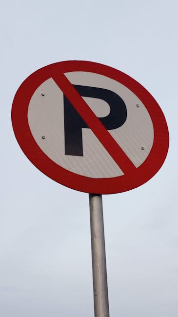 Close-up view of a tall yellow pole supporting a traffic light and two road signs under an overhanging structure. The upper sign displays a black and white speed limit of 10 miles per hour, indicating a low-speed zone likely related to residential or moving areas. Below it, a white sign with black and red markings shows a 'No Parking' restriction with a red circle and slash over a letter 'P', accompanied by a left-pointing arrow. The background reveals a partly cloudy sky with a hint of sunlight. The surrounding environment suggests a street or parking area adjacent to a building or service entrance, with visible roof edges and exterior walls. This setup is typical for areas requiring regulated parking and traffic control during home relocations or moving operations, as managed by companies like Man With a Van Barking, especially when securing parking suspensions or complying with council regulations for furniture transport and home removals.
