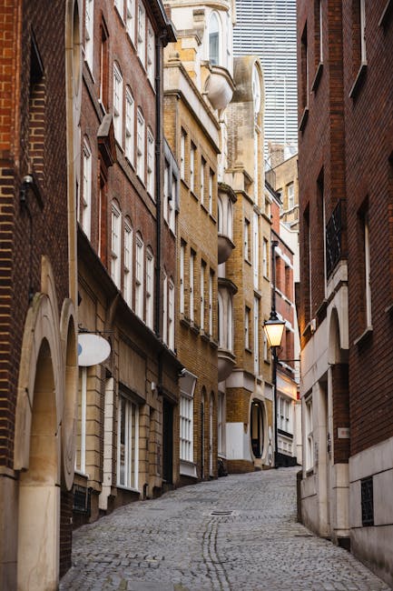 A narrow cobbled street flanked by tall, closely spaced Victorian-style brick buildings with multiple stories and large sash windows, some with decorative stonework or brick detailing. The street gently slopes upward, with a traditional street lamp mounted on the right side, illuminating the area. There are no visible vehicles or pedestrians in the scene, which is taken during daylight with soft, natural lighting. The environment suggests an urban residential area suitable for a home relocation or furniture transport process, consistent with house removals services like those provided by Man With a Van Barking. This setting exemplifies the tight lane conditions encountered during moving projects in the Gascoigne Estate IG11 area, emphasizing the need for careful planning in furniture transport and packing in narrow lanes.