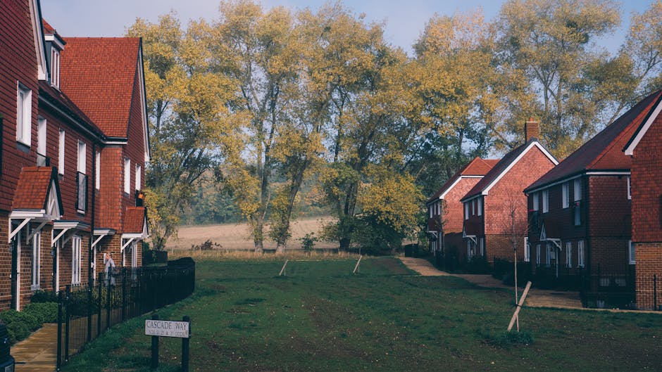 A quiet residential street in Gascoigne Estate IG11 during daytime, featuring neatly arranged red-brick houses with pitched roofs and white-framed windows situated on either side of a grassy pathway. The pathway is partially paved and bordered by black metal fencing along the front gardens of the houses. Several small, newly planted trees with support stakes are visible in the grass, indicating recent landscaping. In the background, tall trees with yellowing autumn leaves create a natural backdrop against a partly cloudy sky, suggesting fall season. The scene is well-lit by natural daylight, highlighting the orderly environment that facilitates house removals and furniture transport. The image reflects a calm, suburban setting often associated with home relocation and logistics by professionals such as Man With a Van Barking, who specialise in removals and moving services, particularly in navigating narrow lanes and residential areas like this.