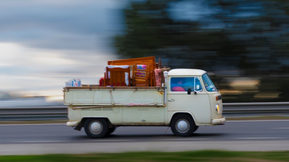 A vintage cream-colored van in motion on a road during daytime, with a blurred background indicating speed. The van's cargo area is loaded with various items, including wooden dining chairs, cardboard boxes, plastic packaging, and bags, all secured for transport. The scene showcases the vehicle's role in home relocation or furniture transport, with a focus on moving logistics such as loading and packing. The setting appears to be outdoor, with the van prominently centered and the motion blur emphasizing its movement, exemplifying a quick and efficient moving service by Man With a Van Barking as indicated on the webpage for last-minute moves in Barking.