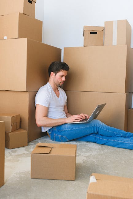 A young man with short dark hair and light skin, wearing a white t-shirt and blue jeans, is sitting on a concrete floor inside a room surrounded by various cardboard boxes, some large and some small, 