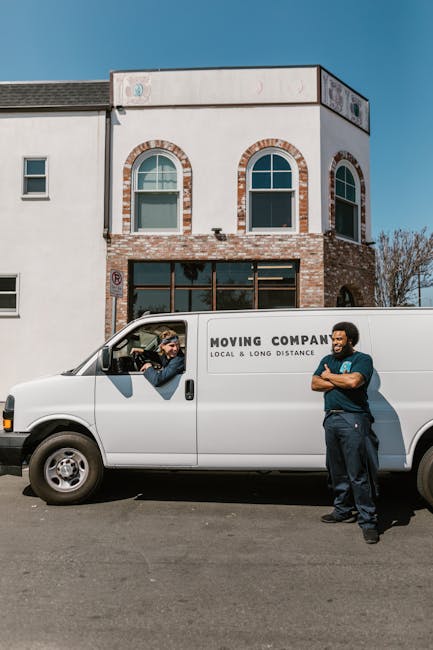 A white moving van from Man With a Van Barking is parked in front of a two-storey residential building with a white and brick exterior, featuring arched windows on the upper floor. The van has the com