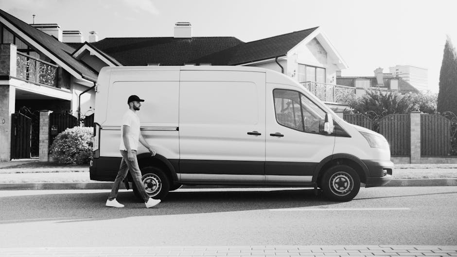 A white moving van from Man With a Van Barking is parked in front of a two-storey residential building with a white and brick exterior, featuring arched windows on the upper floor. The van has the com