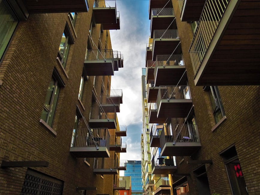 View looking up between two multi-storey residential buildings with brick facades and metal balconies, some with visible furniture or plants. The sky is partly cloudy, and the narrow gap emphasizes the confined space often encountered in house relocation or furniture transport within urban environments. The image captures the external environment where moving services, such as those offered by Man With a Van Barking, may navigate tight terraces and narrow turns involved in home relocations or apartment moves, highlighting logistical challenges faced during loading or unloading processes in limited outdoor spaces.